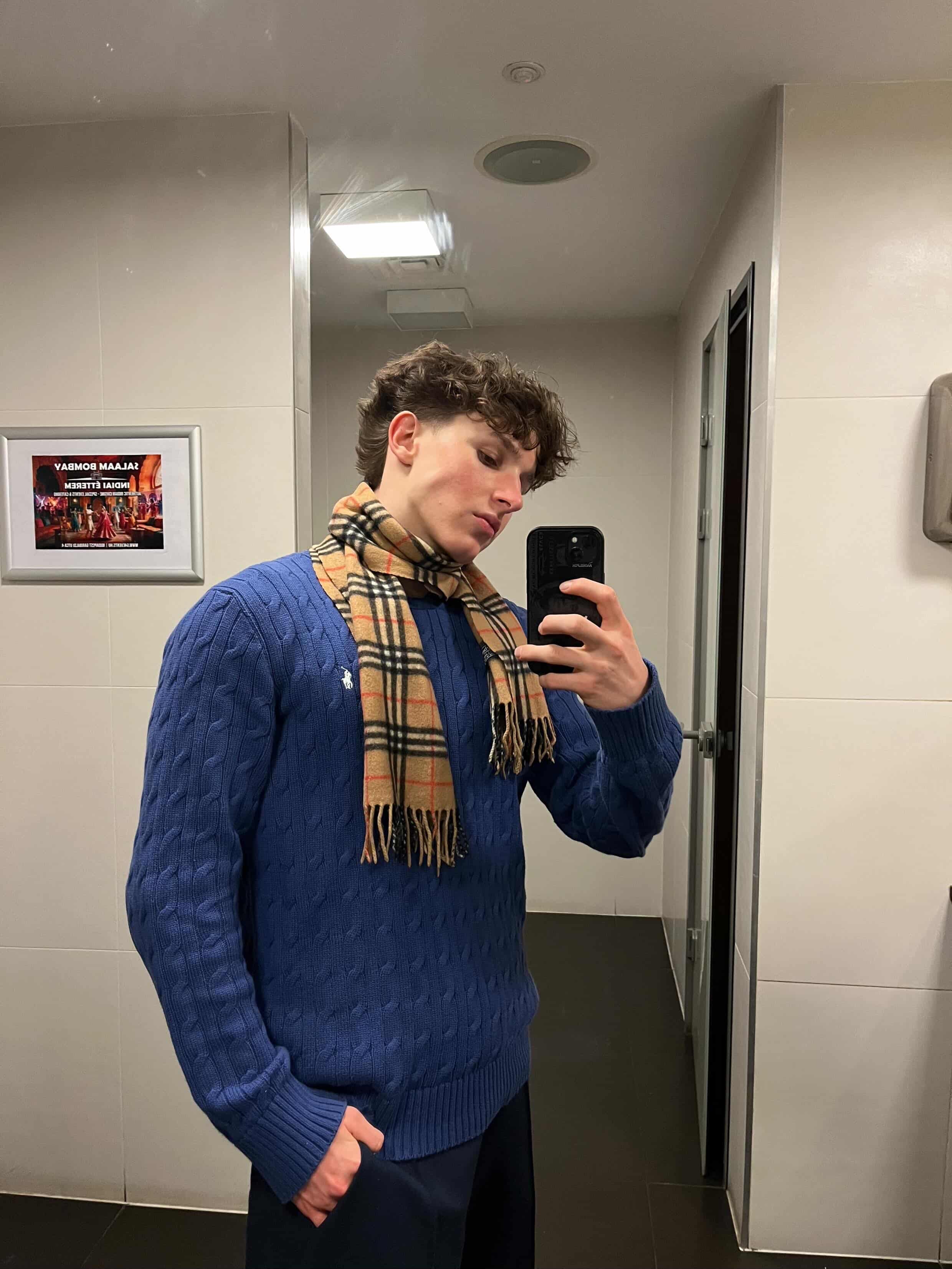 Fiatal férfi tükörszelfin, kék kötött pulóverben és kockás sálban, modern streetwear stílusban.